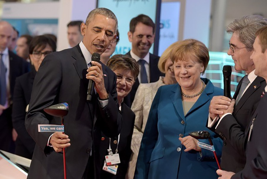 Ex-Präsident Barack Obama zusammen mit Ex-Bundeskanzlerin Angela Merkel und anderen Menschen in Business-Outfits, stehen bei der Hannover-Messe 2010 beieinander und lachen. Obama hält ein Mikrofon und einen Golf-Club in der Hand, auf dem steht "Yes we can"