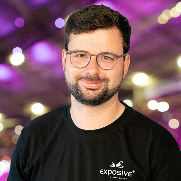 Portraitfoto von Jasper Bitter, Projektleiter bei exposive. Er hat kurze schwarze Haare und einen Vollbart und trägt eine Brille. Er ist bekleidet mit einem schwarzen Shirt mit weißem Branding der exposive medien gruppe. Er lächelt freundlich in die Kamera. Im Hintergrund sieht man ein Foto mit starkem Bokeh-Effekt, vom Stadionsingen in Hannover. Die vorherrschenden Farben sind violett und grün. Viele Lichter erstrahlen in warmweiß.