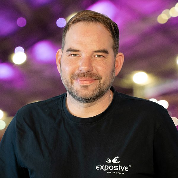 Portraitfoto von Thomas Lux, Projektleiter bei exposive. Er hat kurze dunkle Haare und lächelt freundlich in die Kamera. Er ist bekleidet mit einem schwarzen Shirt mit weißem Branding der exposive medien gruppe. Im Hintergrund sieht man ein Foto mit starkem Bokeh-Effekt, vom Stadionsingen in Hannover. Die vorherrschenden Farben sind violett und grün. Viele Lichter erstrahlen in warmweiß.